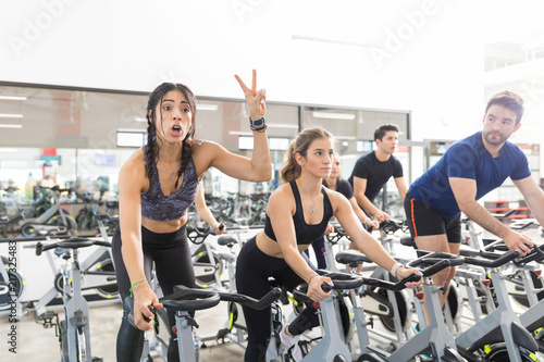 Fototapeta Naklejka Na Ścianę i Meble -  Woman Gesturing Victory While Exercising On Spinning Bike In Gym