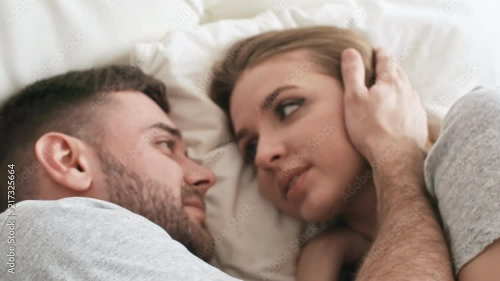 Handheld shot with close up of affectionate bearded man lying in bed and stroking face of beautiful young woman smiling and talking while looking into his eyes Handheld shot with close up of affectionate bearded man lying in bed and stroking face of beautiful young woman smiling and talking while looking into his eyes