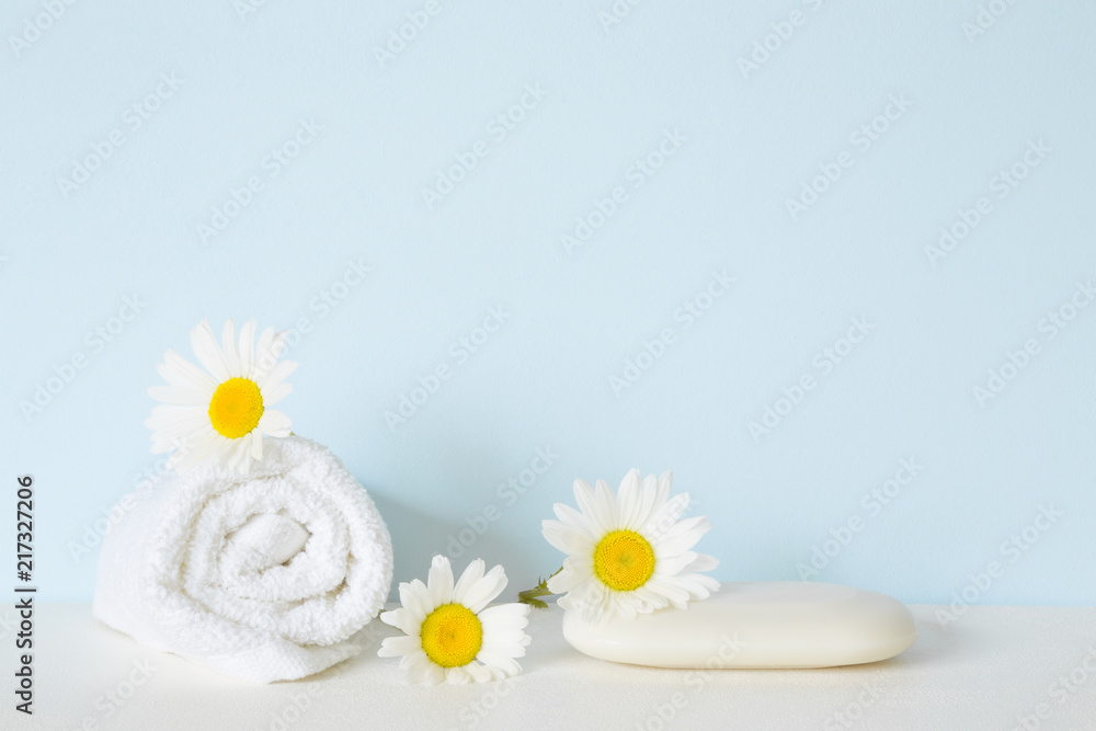 White natural soap and towel on shelf in bathroom. Beautiful camomiles or daisies. Fresh flowers. Care about clean and soft face, hands, legs and body skin. Empty place for text on pastel blue wall. White natural soap and towel on shelf in bathroom. Beautiful camomiles or daisies. Fresh flowers. Care about clean and soft face, hands, legs and body skin. Empty place for text on pastel blue wall.