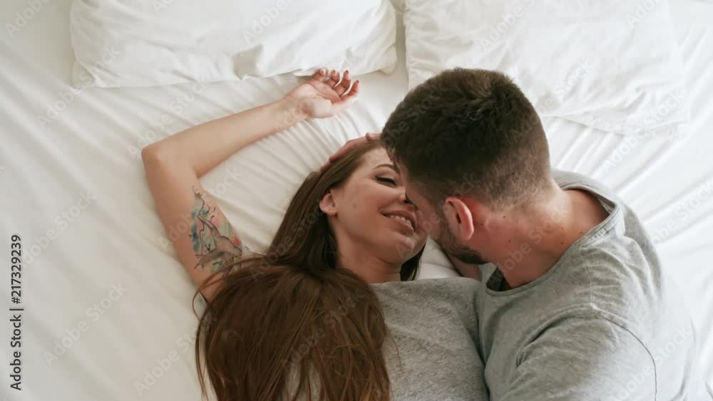 Handheld top view shot of happy young woman lying down on soft bed and laughing as her bearded boyfriend kissing her on lips Handheld top view shot of happy young woman lying down on soft bed and laughing as her bearded boyfriend kissing her on lips
