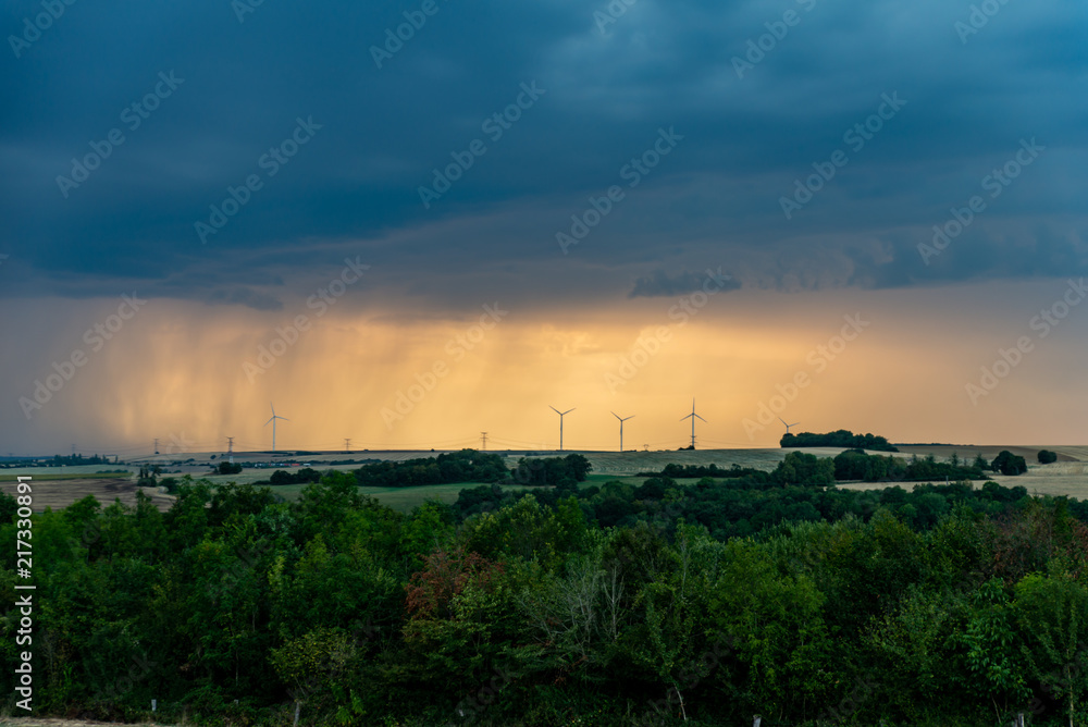 Paysage de campagne au crépuscule orange, l'orage se prépare à éclater, la pluie au loin lazarde le ciel au dessus de l'horizon et des éoliennes