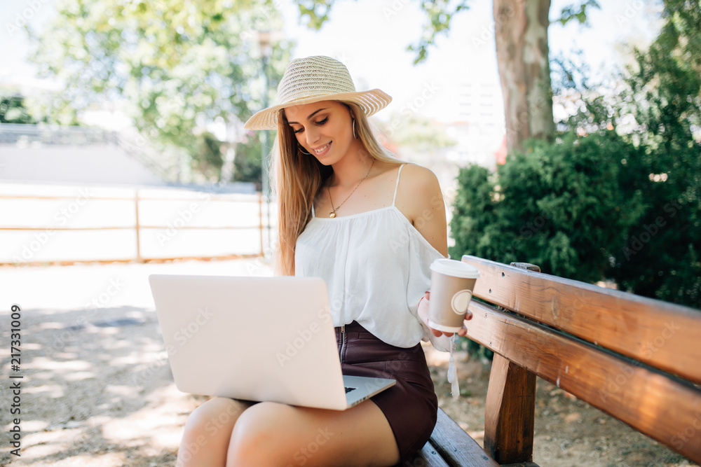 Portrait of attractive young woman sitting on bench in green park on summer day and using silver laptop while drinking takeaway coffee from paper cup