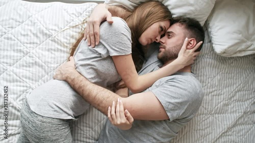 Handheld top view of affectionate young woman and bearded man lying on bed and hugging: they are looking into each others eyes and sharing intimate moment