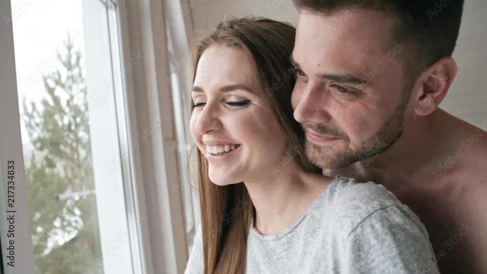 Handheld shot of cheerful bare-chested man with beard hugging happy young girlfriend and talking to her while looking out window Handheld shot of cheerful bare-chested man with beard hugging happy young girlfriend and talking to her while looking out window