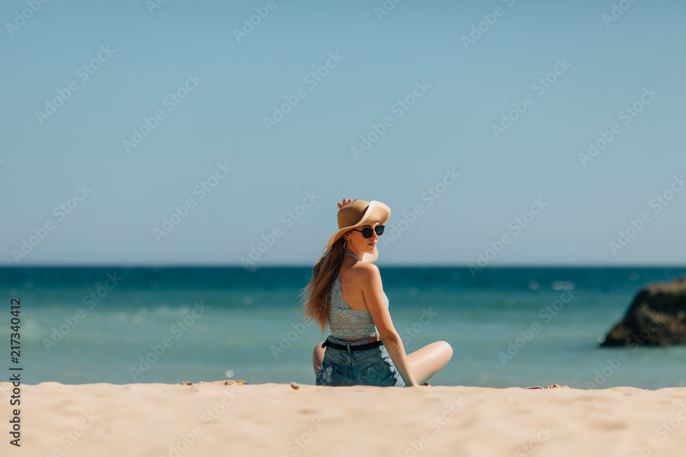 Rear view of a young woman with straw hat looking at an idyllic view while sitting on the sand at ocean beach Rear view of a young woman with straw hat looking at an idyllic view while sitting on the sand at ocean beach