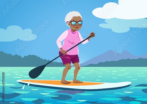 Senior African American woman on a stand up paddle board. Cute grandma wearing rash guard and shorts paddling on calm water, trees and mountains in the background. Healthy active lifestyle for seniors