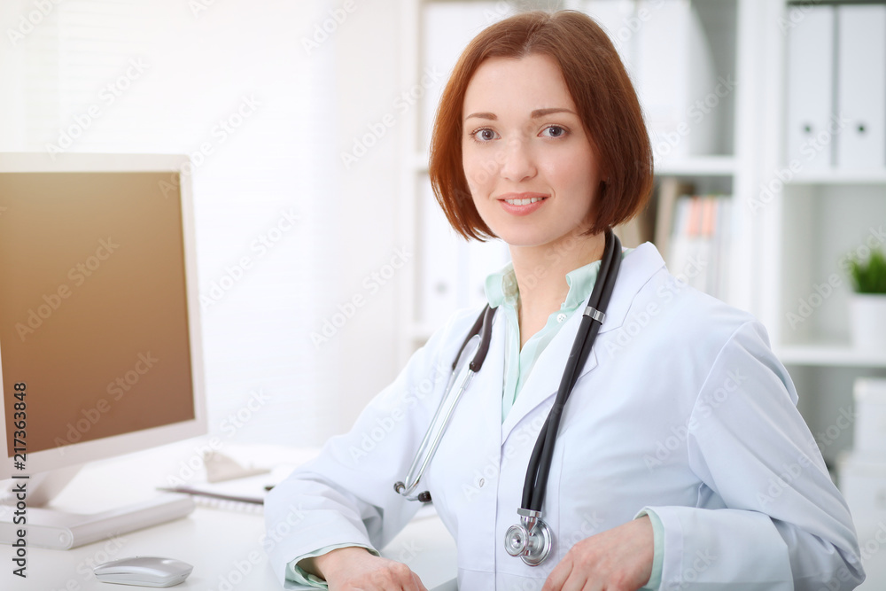 Young brunette female doctor sitting at the table and working with computer at hospital office Young brunette female doctor sitting at the table and working with computer at hospital office