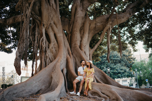 Obraz na płótnie Beautiful young brunette woman with her boyfriend with beard sitting on the large root of the giant tree in Spain in the evening w salonie