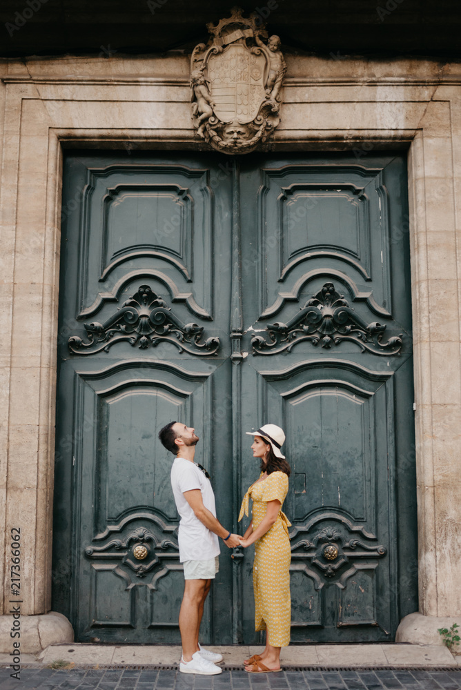 Beautiful young brunette woman with her boyfriend with beard staying holding hands with an old giant historic door on the background in Spain in the evening