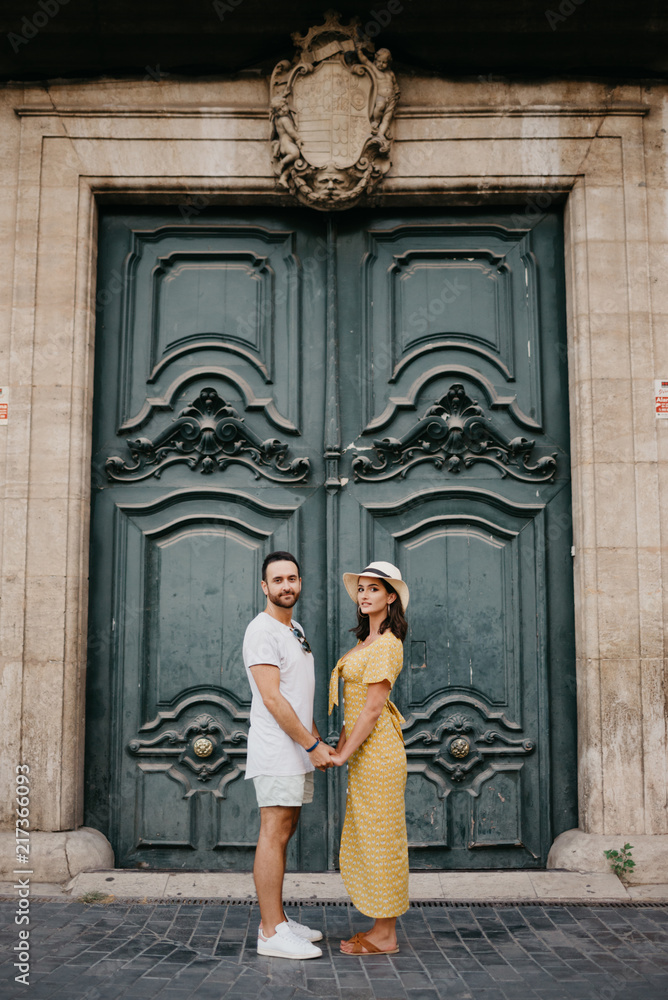 Pretty brunette girl with her boyfriend with beard staying holding hands with an old giant historic door on the background in Spain in the evening