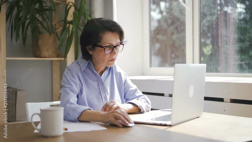 Mature businesswoman is working with laptop sitting in home office space, stylish business lady wearing eyeglasses is writing, typing at table with pc near window in bright room. Concept: work day
