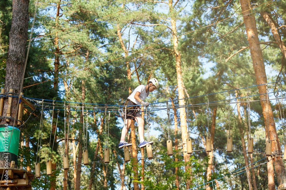 Fototapeta premium cute boy in white t shirt in the adventure activity park with helmet and safety equipment. Young boy playing and having fun doing activities outdoors. Hobby, active lifestyle concept