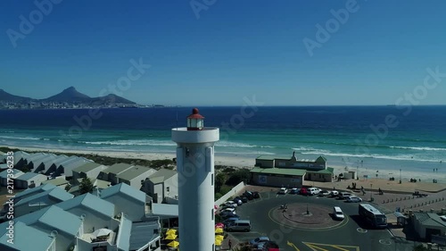 Aerial orbit or point of interest around Milnerton Lighthouse in Cape Town South Africa, revealing white sandy beach with waves crashing through as well as iconic Table Mountain.