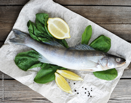 Raw pikeperch on parchment paper. Next to the fish are fresh spinach and lemon slices. Light wooden background. View from above. Close-up.