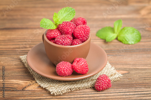 Ripe raspberries with green mint leaves in brown cup and saucer on sackcloth and wooden background.