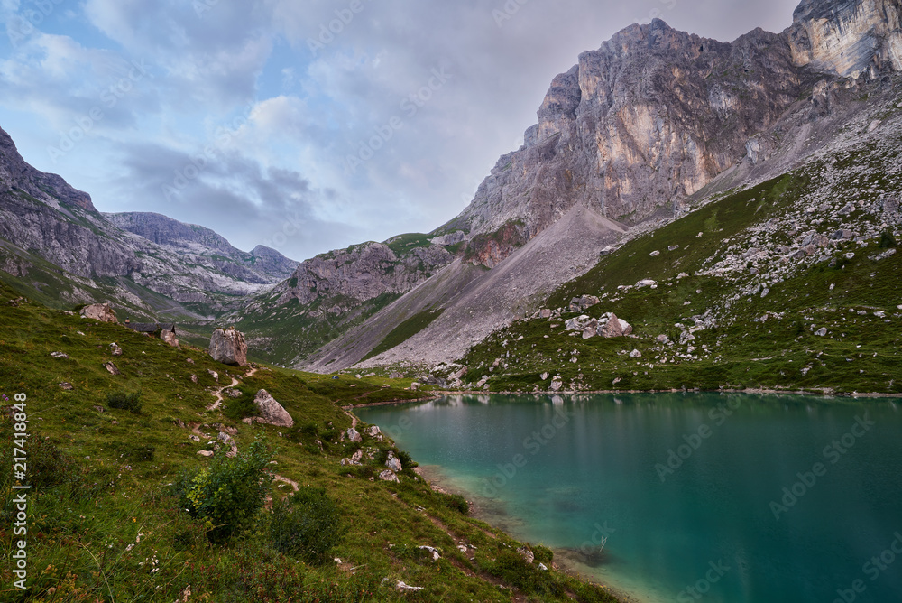 Partnunsee, türkisblauer Bergsee und Gebirgslandschaft im Abendlicht, blauer Himmel mit Wolken, schroffe Felsen, Alpwiese mit Wanderweg