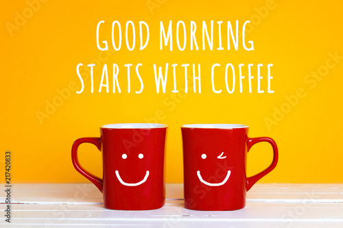 Two red coffee mugs with a smiling faces on a yellow background with the phrase Good morning start with coffee.