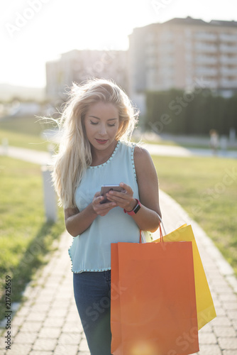 Woman walking in park with shopping bags and looking smartphone while sending message photo