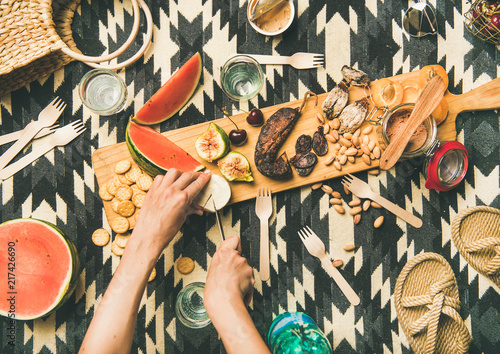 Summer picnic setting. Flat-lay of fresh fruit, smoked sausage, nuts, pate, cracker, straw accessories and woman hands cutting brie cheese over linen blanket, top view. Outdoor gathering or lunch