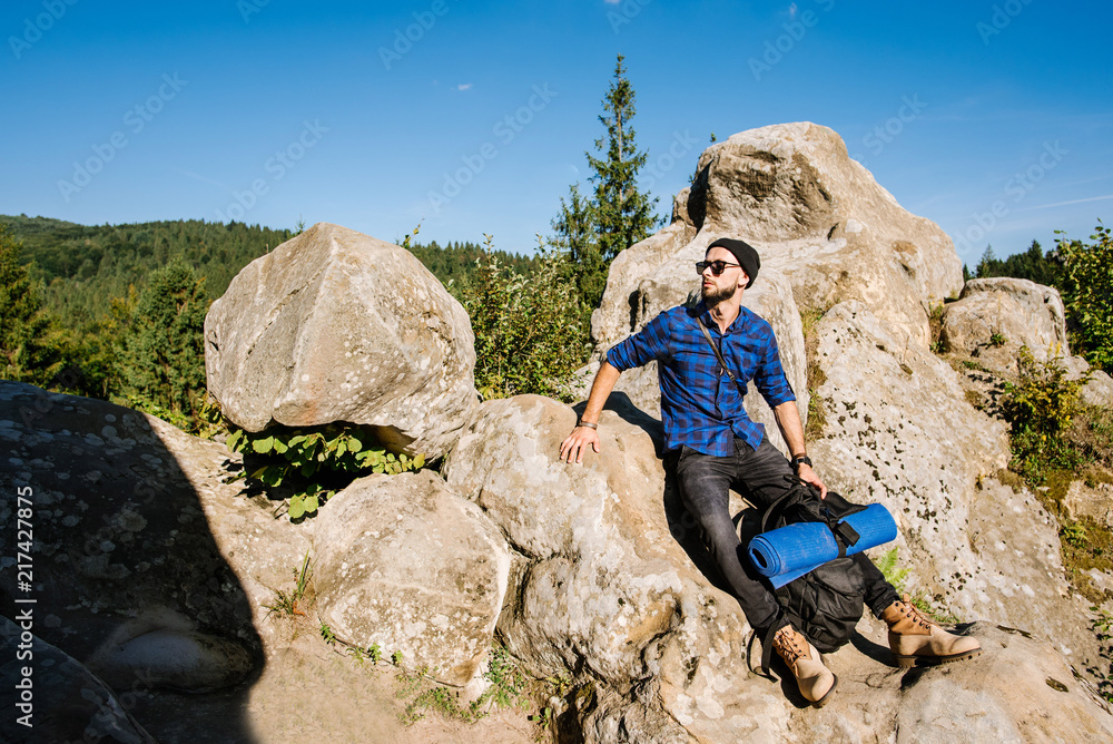 A young man traveler with backpack wearing sunglasses sitting on the top of rocky mountains