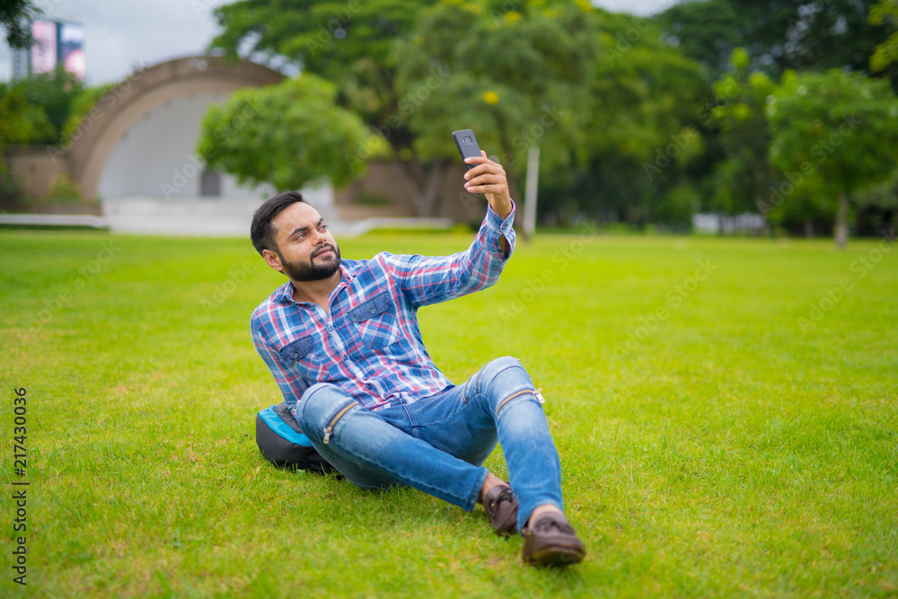 Handsome Indian Man In Park Lying Down On Grass And Taking Selfie With Mobile Phone Handsome Indian Man In Park Lying Down On Grass And Taking Selfie With Mobile Phone