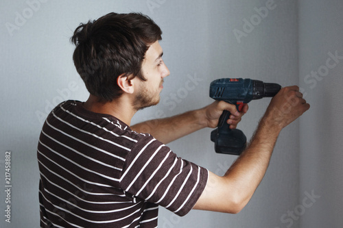 Young handsome guy with a screwdriver is drilling a white wall in a new apartment doing repairs.