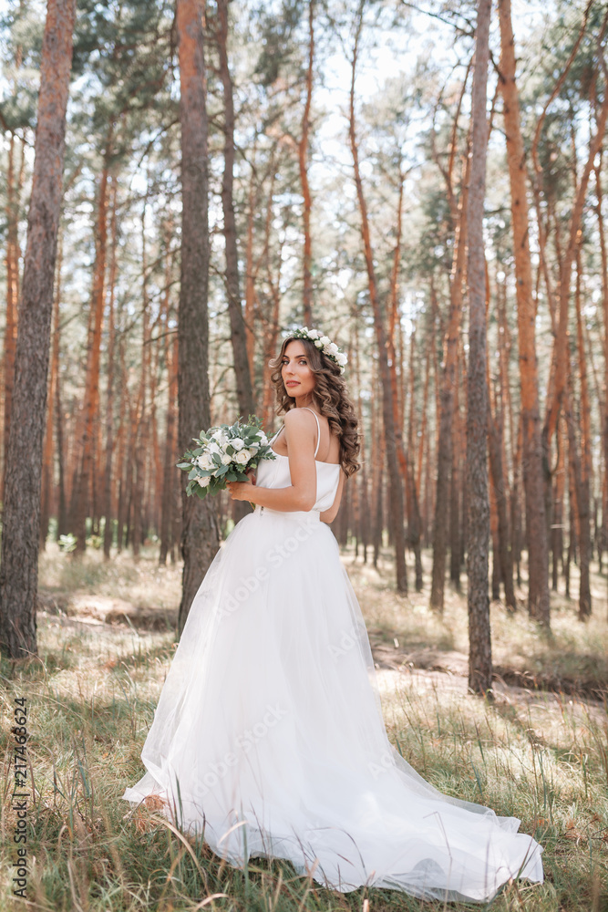 A cute curly woman in a white wedding dress with a wedding bouquet and wreath in her hair standing back to the camera in nature. Concept escaped bride. Forward to a happy bright future Runaway