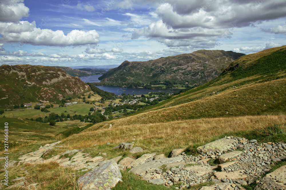 Naklejka premium Landscape scene of Ullswater, one of the largest lakes in the English Lake District, and the village of Glenridding, taken from Birkhouse Moor and set against a brilliant blue summer sky with white cl