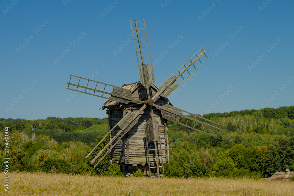 Antique old wooden windmill under blue sky  in front of forest. Sunny summer day. National Museum of Folk Architecture and Life of Ukraine in Pyrohiv. 