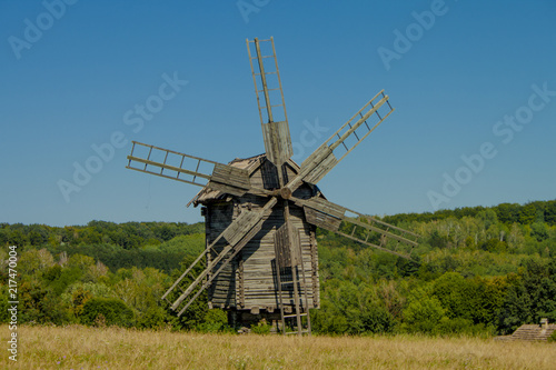 Antique old wooden windmill under blue sky in front of forest. Sunny summer day. National Museum of Folk Architecture and Life of Ukraine in Pyrohiv. 