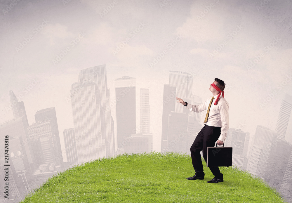 Young blindfolded businessman steps on a a patch of grass with a cloudy city in the background Young blindfolded businessman steps on a a patch of grass with a cloudy city in the background