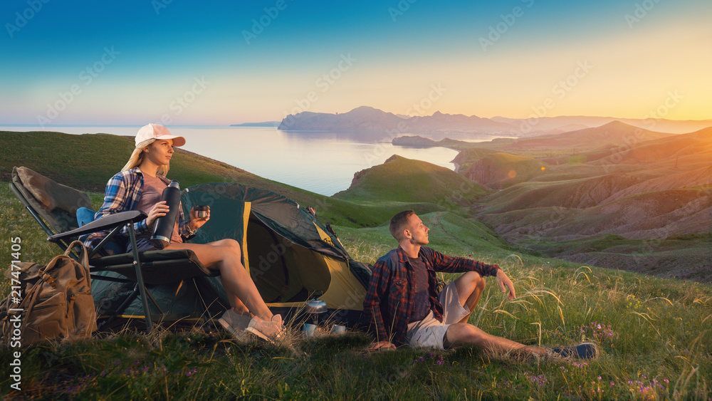 Fototapeta premium Happy backpackers with a tent look at the sunset in mountains. Young couple hikers enjoys the sunset and nature. People in camping. Adventure hiking trip. Romantic journey.