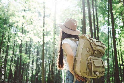 Back view on cute young woman with hat, backpack and location map in hand among trees in forest at sunset.