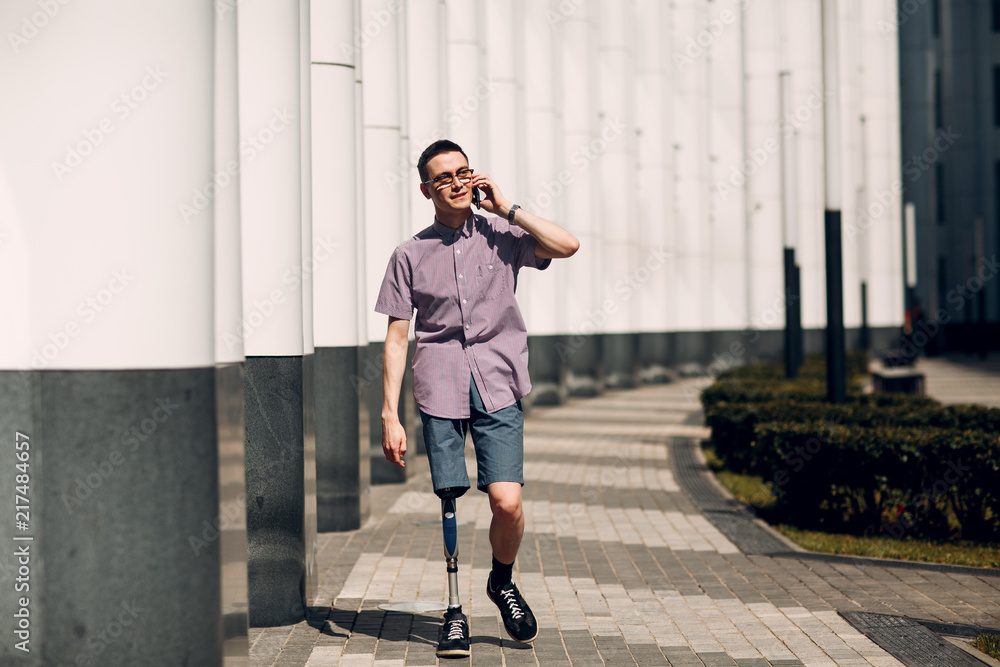 Disabled young man with foot prosthesis walks along the street and hold mobile phone