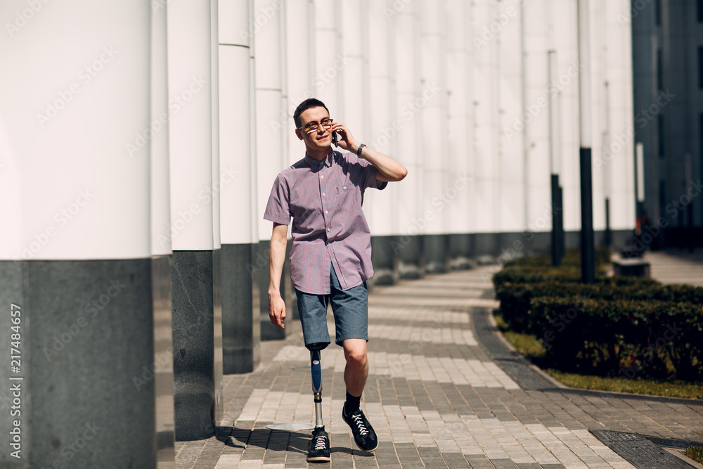 Disabled young man with foot prosthesis walks along the street and hold mobile phone Disabled young man with foot prosthesis walks along the street and hold mobile phone