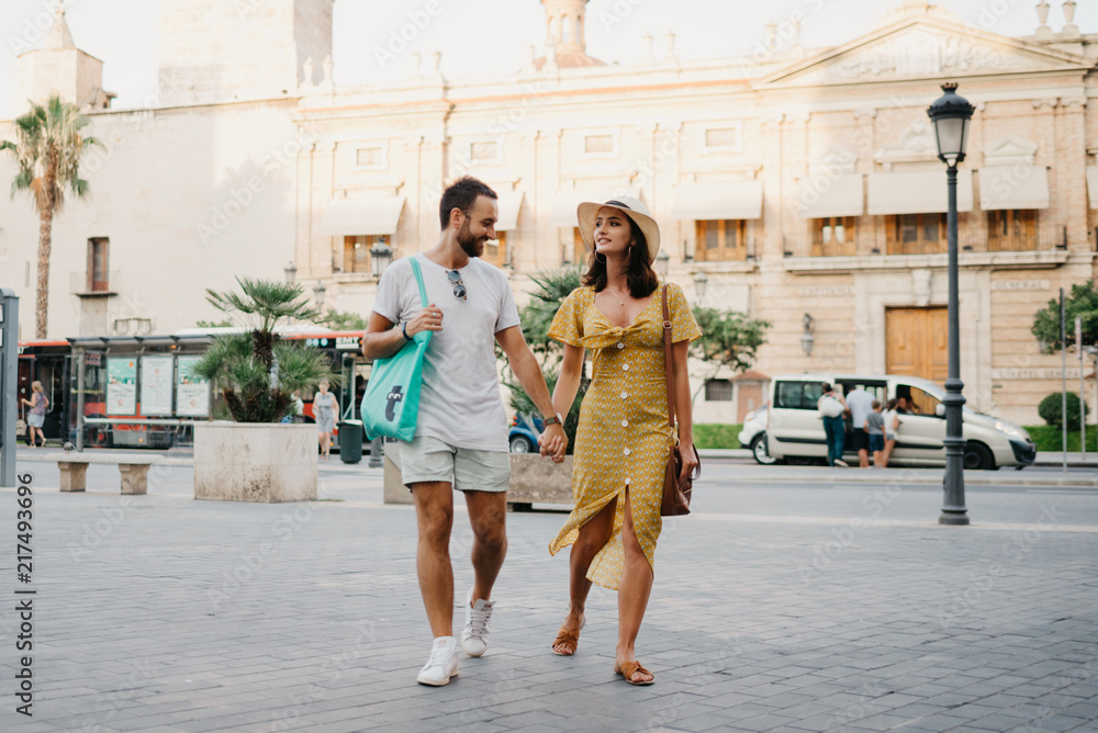 Awesome brunette girl in the hat with her boyfriend with beard walking together on the old European street in Spain on the sunset