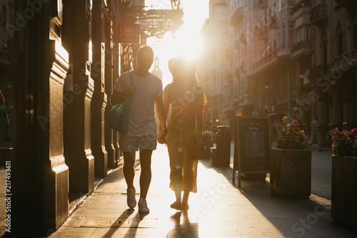 Awesome girl with hat with her boyfriend with beard walking together holding hands in the orange rays of the sunset on the old European street in Spain on the sunset