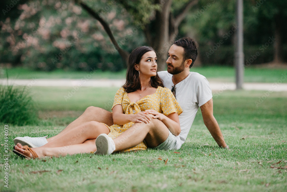 Stylish cute brunette girl sitting between legs of her boyfriend with beard  close to each other on the grass in the park in Spain in the evening Stock  Photo