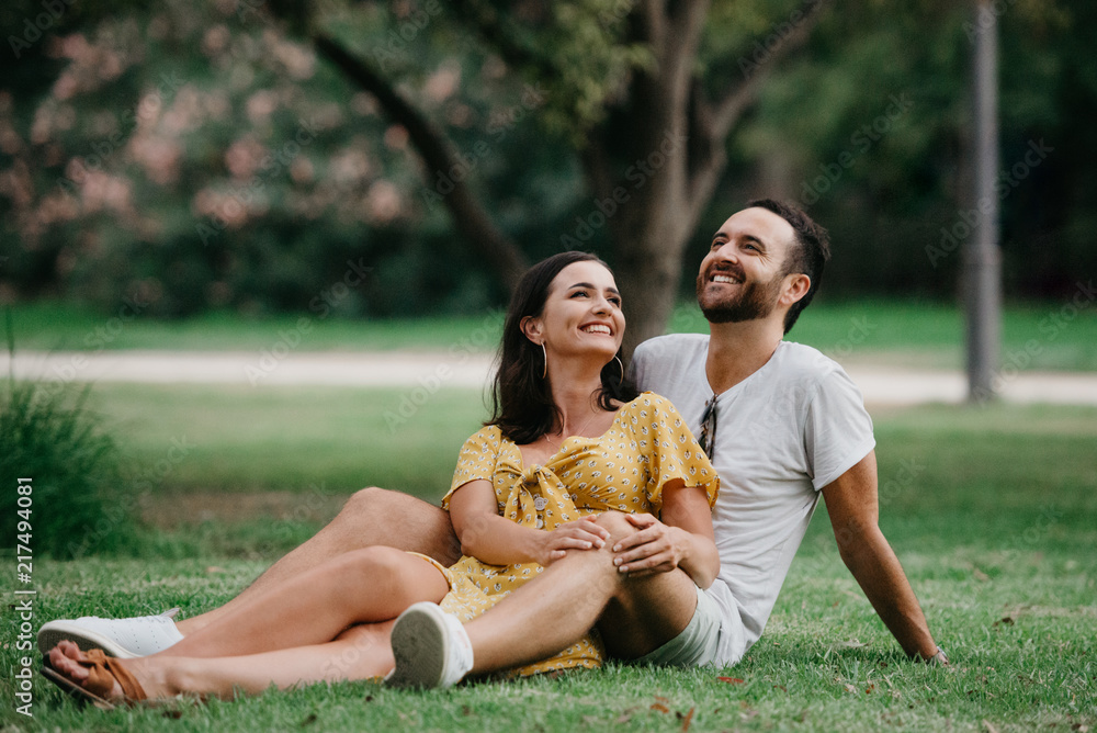 Stylish cute brunette girl sitting between legs of her boyfriend with beard  close to each other on the grass in the park in Spain in the evening, and  they both laughing Stock