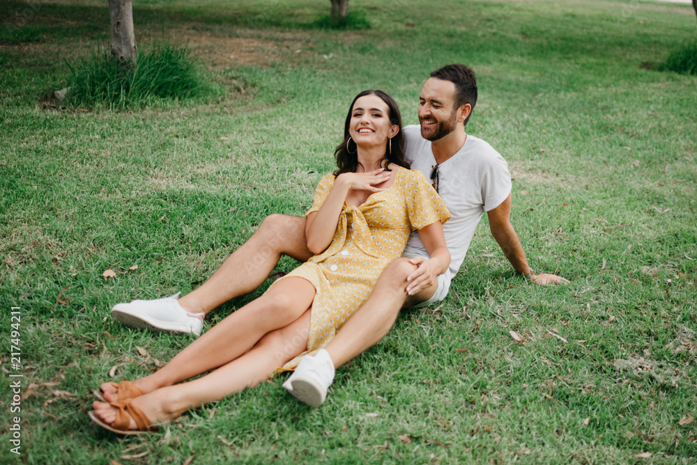 Foto de Stylish cute brunette young girl laughing sitting between legs of  her boyfriend with beard close to each other on the grass in the park in  Spain in the evening do