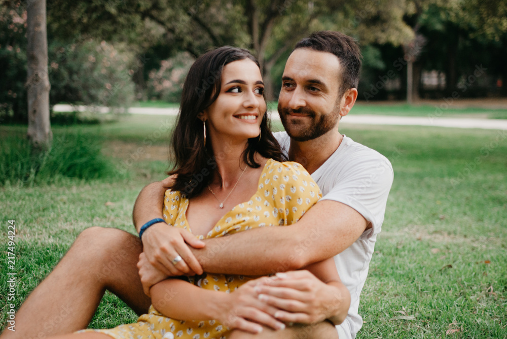 Naklejka premium Stylish young man with beard hugging his cute smiling girlfriend sitting between his legs on the grass in the park in Spain in the evening