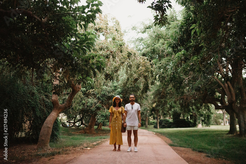 Stylish cute brunette girl with her boyfriend with beard posing holding hands close to each other on the small road in the park between large trees in Spain in the evening. Date of the young couple