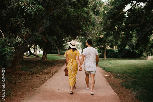 Stylish cute brunette girl with her boyfriend with beard walking holding hands close to each other on the small road in the park between large trees in Spain in the evening. Date of the young couple