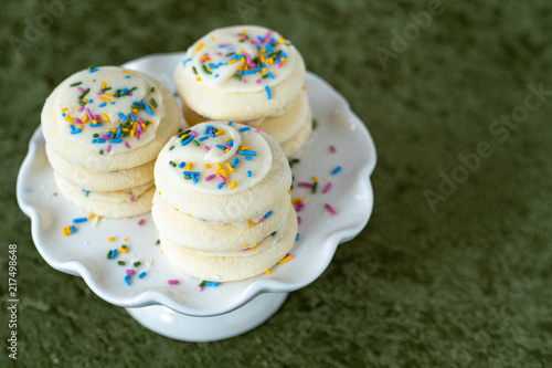 Three stacks of vanilla frosted sugar cookies with multi-color sprinkles on a white cake plate, on a green background
