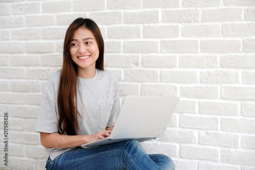 Young asian woman using laptop computer sitting in front of white brick wall background with copy space, people and technology, lifestyles, education, business concept