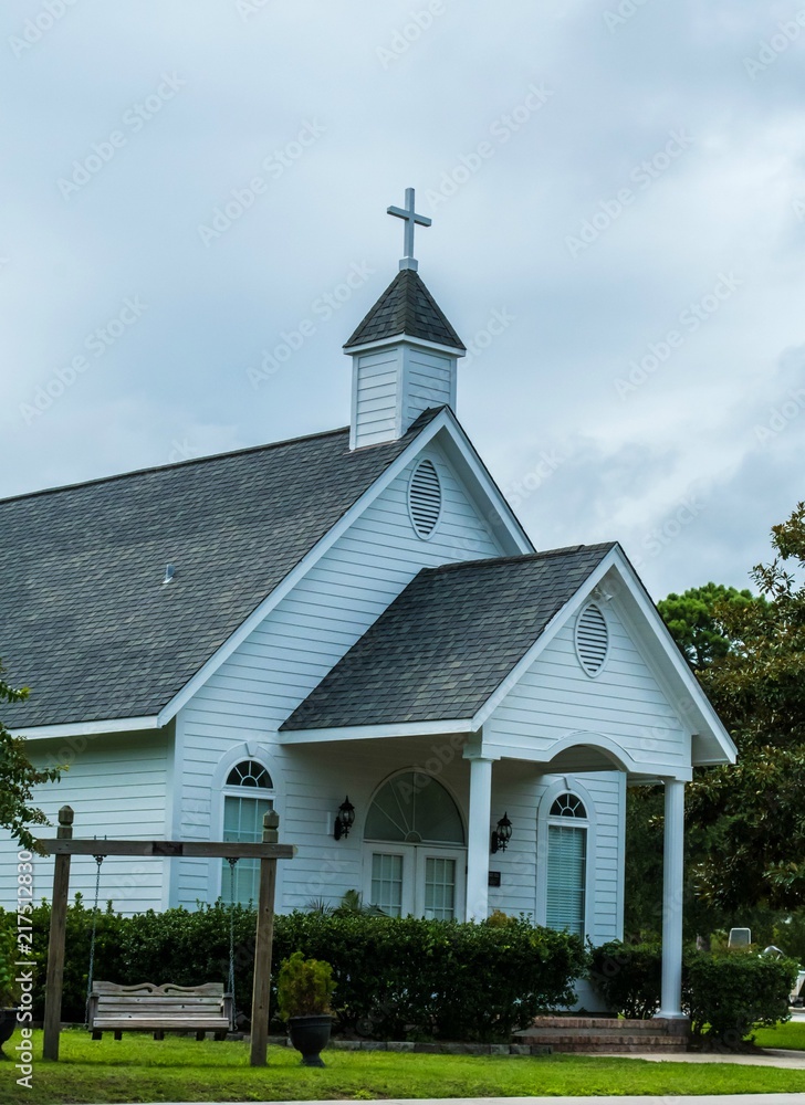 White country church with wooden siding, swing bench, old wooden cross on the cupola, vertical orientation, symbol of religion and faith, as dark grey clouds loom above