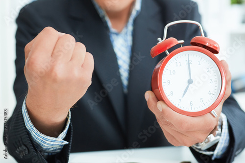 Young man in suit showing an alarm clock set at seven and fist. Boss is angry at the late arrival of employees. photo