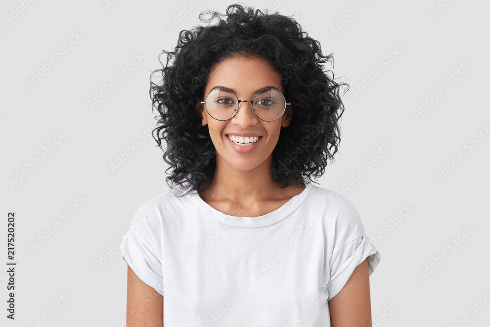 © wayhome.studio  - Beautiful smiling African American female with crisp hair, broad smile, shows white teeth, wears casual t shirt and spectacles, stands over studio wall rejoices having day off. Woman journalist indoor