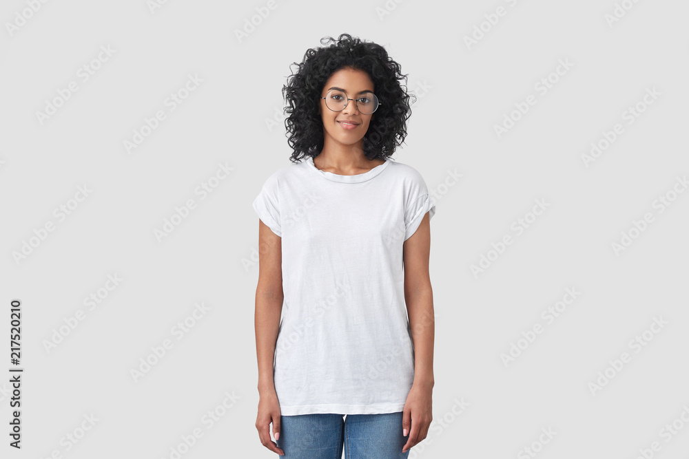 Fotografija Indoor shot of attractive dark skinned female with crisp hair, wears round glasses, dressed in casual white t shirt and jeans, poses indoor, has Afro hairstyle
