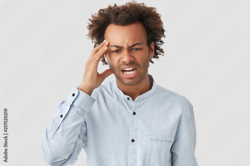 Stressful young African American male keeps hand on temple, looks desperately down, has headache, curly hair, frowns face in discontent, dressed in elegant shirt, isolated over white background Stressful young African American male keeps hand on temple, looks desperately down, has headache, curly hair, frowns face in discontent, dressed in elegant shirt, isolated over white background